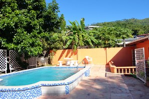 A rectangular swimming pool with blue mosaic tiles, surrounded by a patio area with a table and chairs, and a fence.