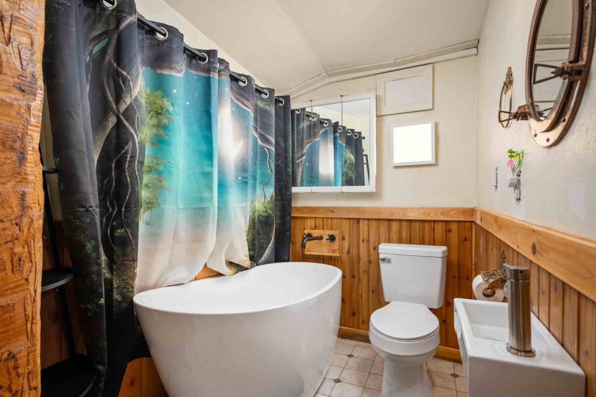 Une salle de bain avec un mur recouvert de panneaux en bois, une baignoire blanche, des toilettes blanches et un rideau de douche orné d’un motif représentant un paysage naturel.