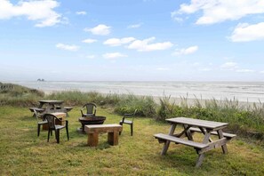 Un área de picnic con bancos y sillas de madera, con vista a una playa con olas y accidentes geográficos lejanos.