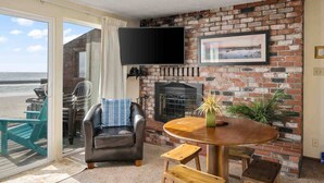 A living room with a brick fireplace, a flat-screen TV mounted above it, and a view of the beach through a sliding glass door.