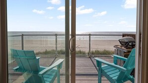 A balcony with two turquoise chairs and a grill, overlooking a beach with a clear sky.