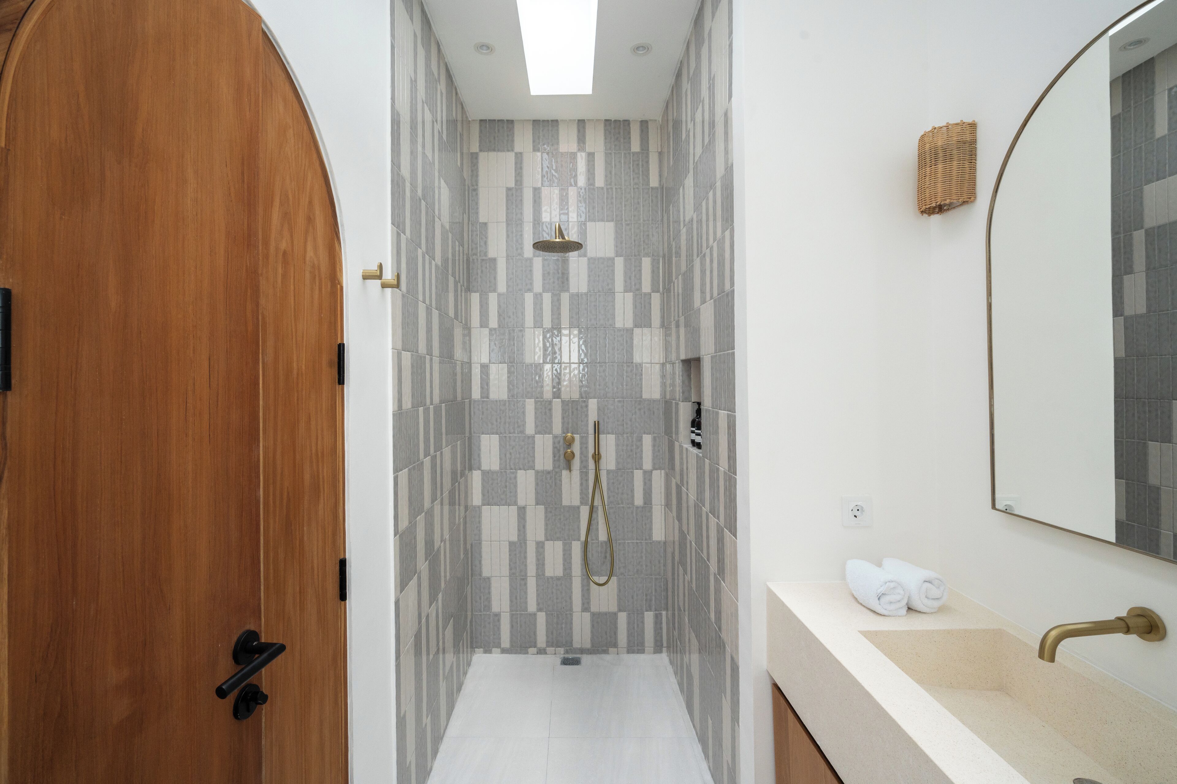 A modern bathroom with a wooden door, a shower with a gold showerhead, a white sink, and a mirror.