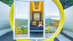 A circular, yellow-themed room with a bed, pillows, and a window overlooking a scenic landscape. - The Hosteller Panshet, Lakeview  (Pune)