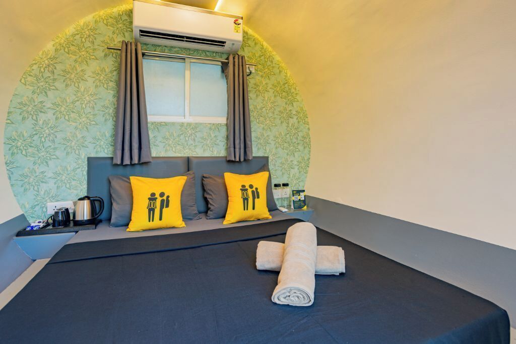 A neatly made bed with a rolled towel, a window with curtains, and a wall with a floral pattern.