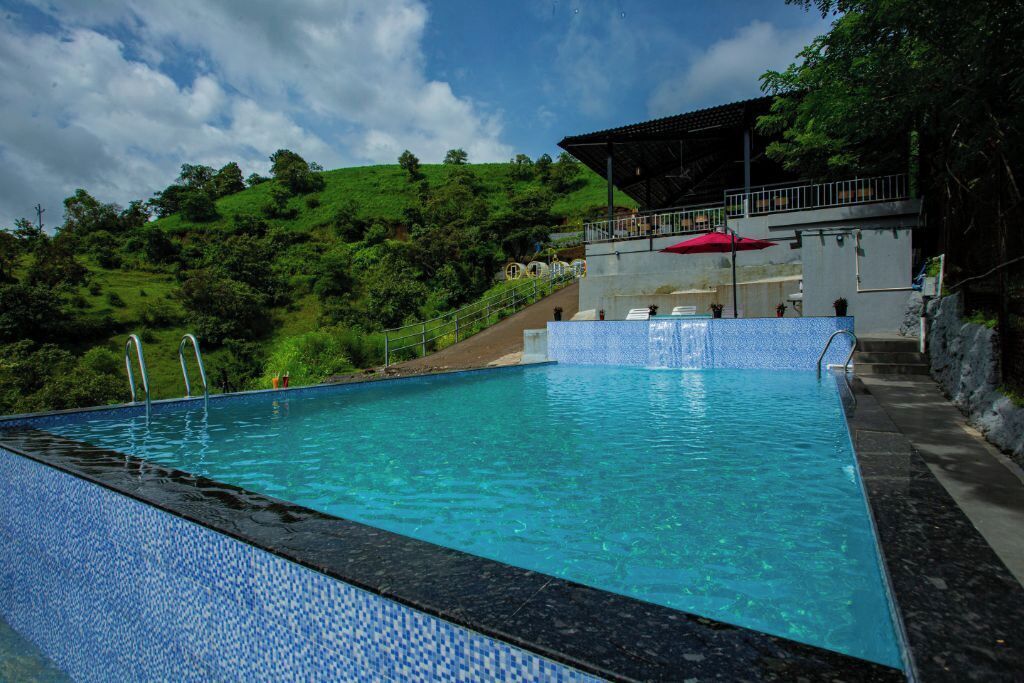 A swimming pool with a tiled edge, a covered seating area with a red umbrella, and a hillside in the background.