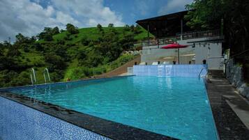 A swimming pool with a tiled edge, a covered seating area with a red umbrella, and a hillside in the background.