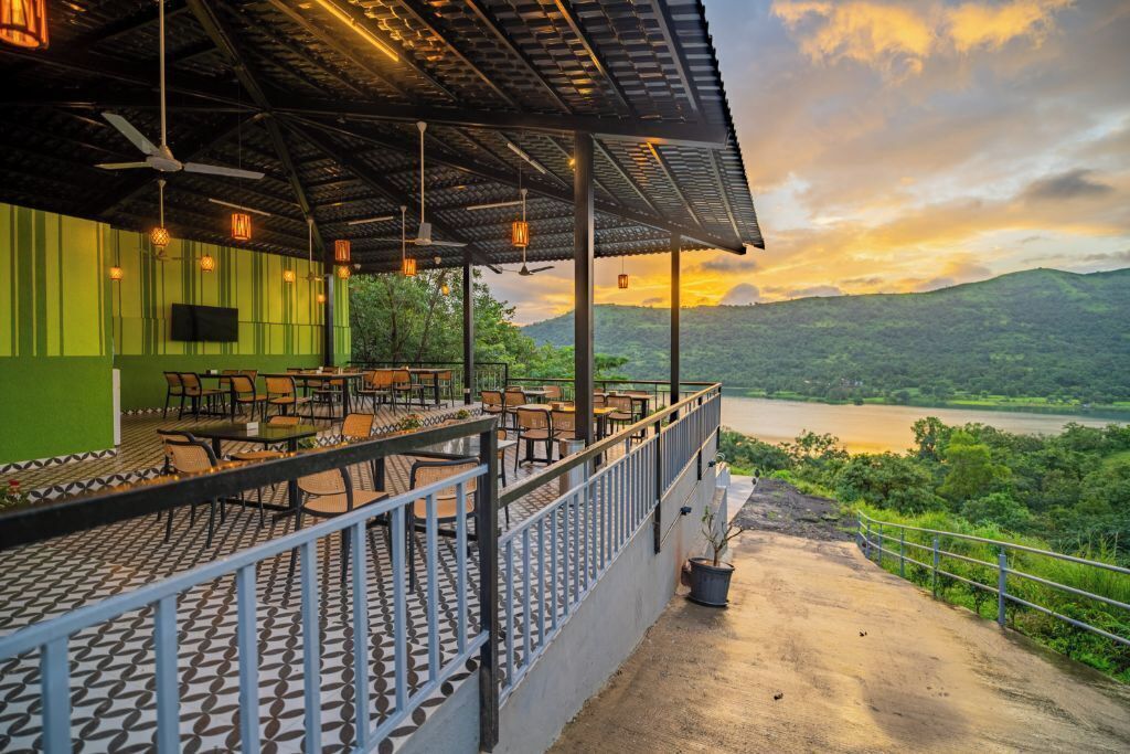 An outdoor restaurant with a terrace overlooking a river, featuring tables and chairs, a green wall, and a sunset view.