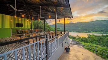 An outdoor restaurant with a terrace overlooking a river, featuring tables and chairs, a green wall, and a sunset view.