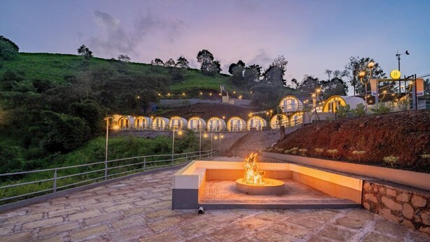 A stone-paved outdoor area with a fire feature and a series of illuminated structures on a hillside. - The Hosteller Panshet, Lakeview  (Pune)
