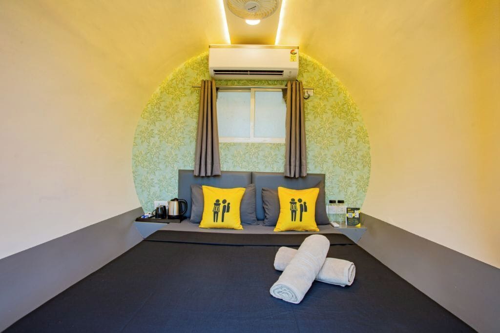 Ein Hotelzimmer mit einem Bett, zwei Kissen mit Symbolen, einer Klimaanlage, einem Fenster mit Vorhängen und einem kleinen Tisch mit Gegenständen.