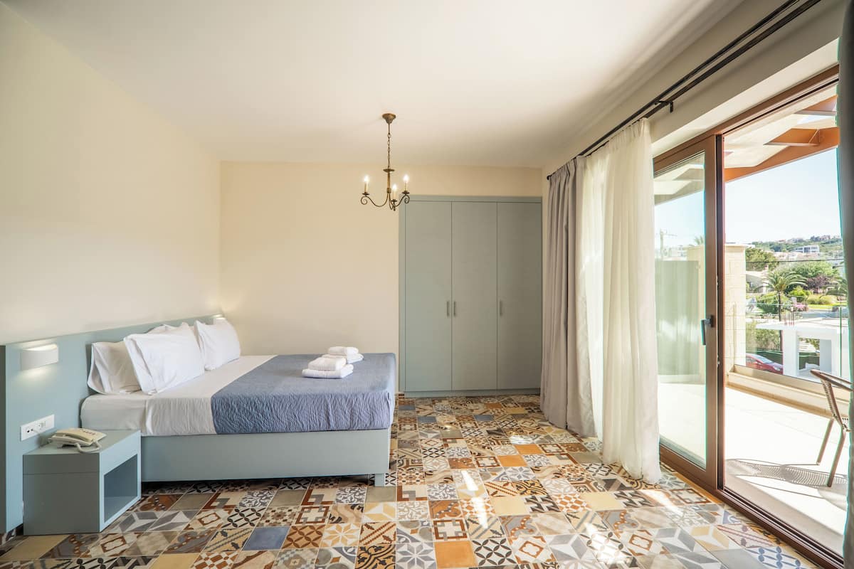 A bedroom with a bed, a nightstand, a wardrobe, a patterned floor, and a view through a sliding glass door.
