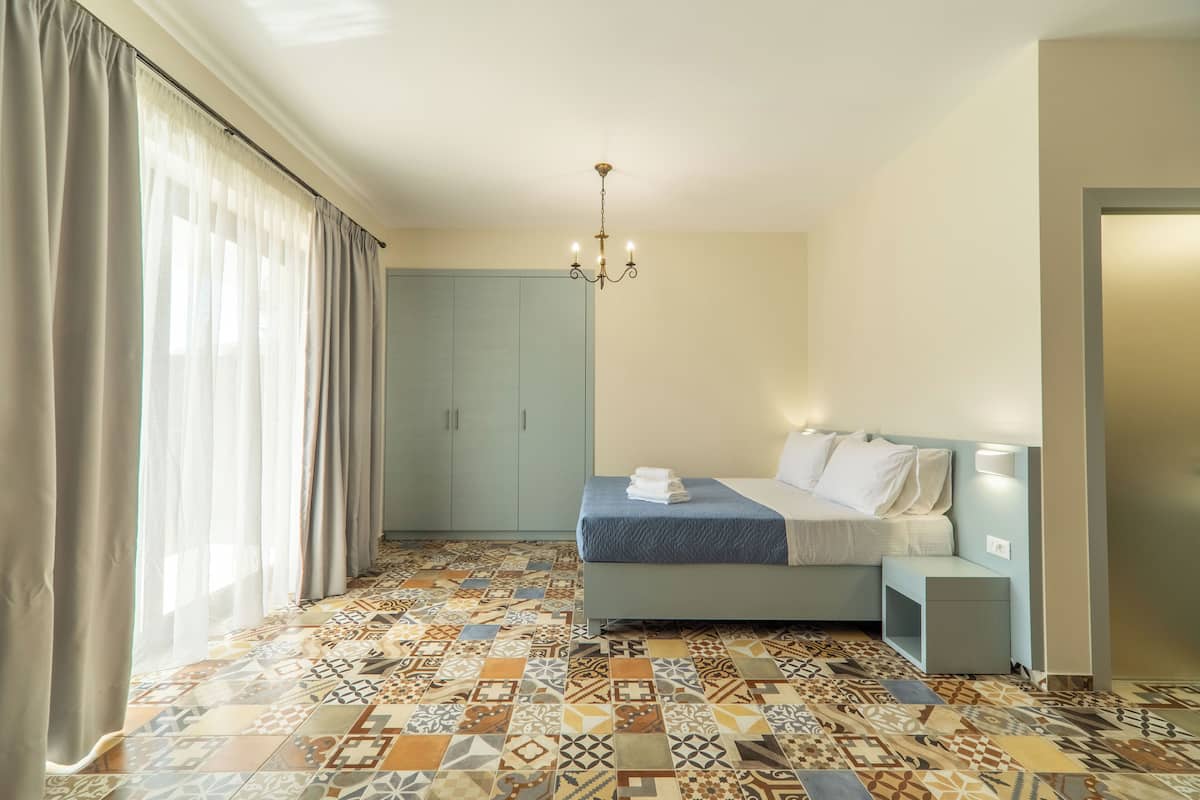 A bedroom with a patterned floor, a bed with a blue cover, a nightstand, a wardrobe, and a chandelier.