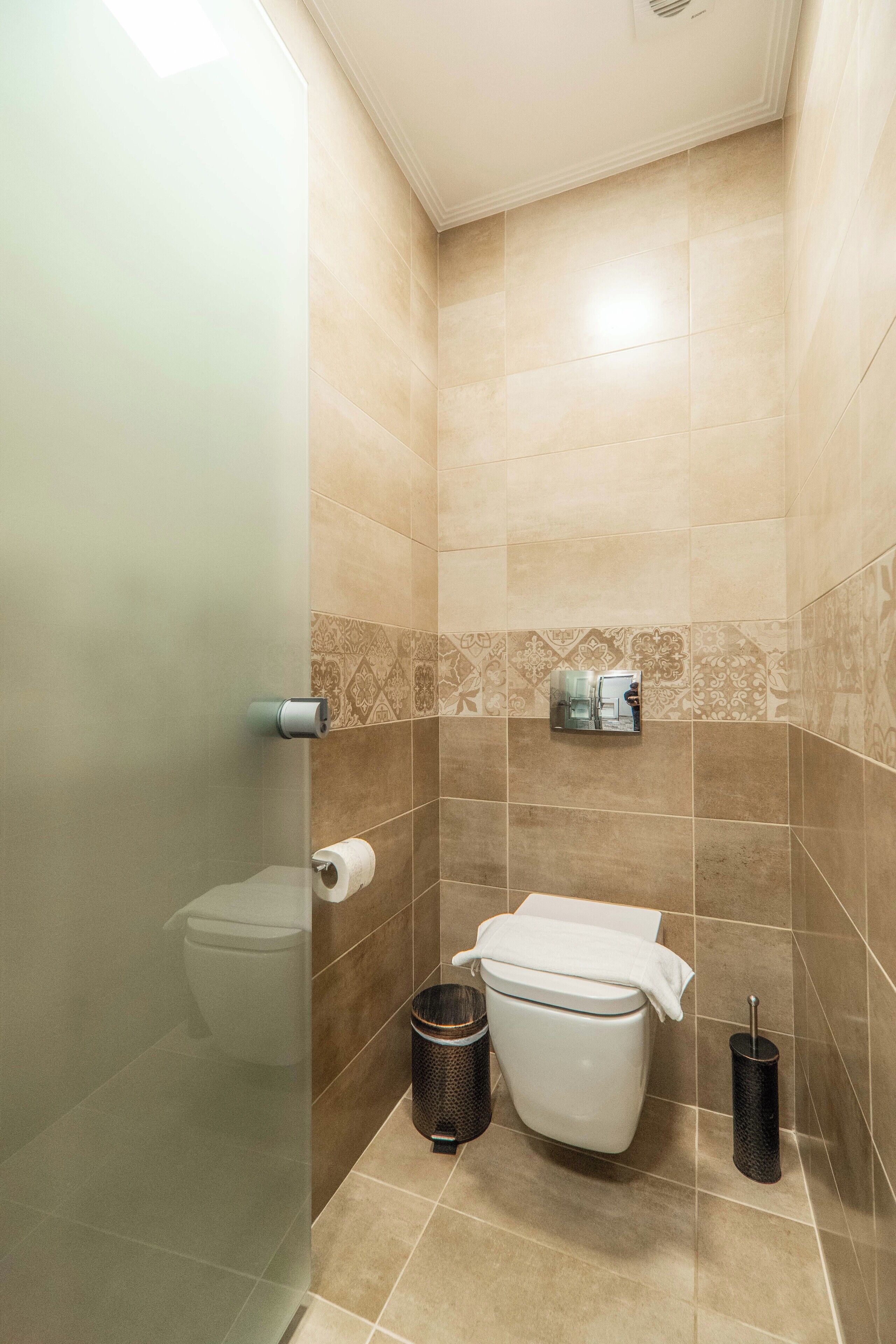 Une salle de bain avec des carreaux de couleur beige, des toilettes, une poubelle et un porte-rouleau de papier toilette.