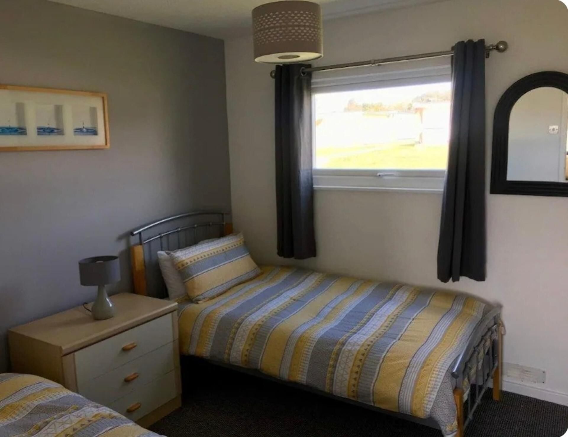 Un dormitorio con cama, una mesita de noche con lámpara, una ventana con cortinas, un espejo y un cuadro en la pared.