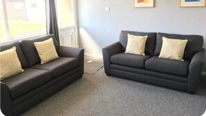 Une pièce avec deux canapés gris foncé, chacun agrémenté d’un coussin de couleur claire, et un tapis gris. Deux œuvres d’art encadrées ornent le mur, et une fenêtre avec des rideaux est visible.