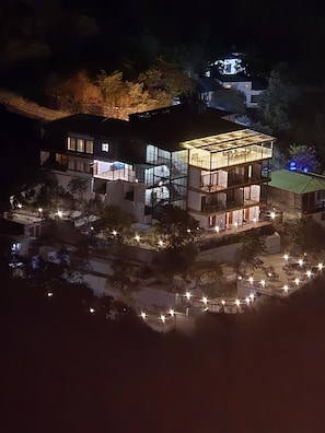Un edificio moderno de varios pisos con ventanas grandes y un área de balcón, rodeado de árboles y un espacio exterior bien iluminado.