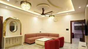 A modern living room with a sectional sofa, a coffee table, and a red ottoman. There is a ceiling fan, chandeliers, and a television mounted on the wall. - Gurugram Resort Mussoorie (Mussoorie)