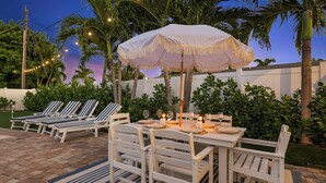 Un espace de restauration en plein air avec des chaises blanches et une table dressée pour un repas, entouré de palmiers et d’une clôture blanche.