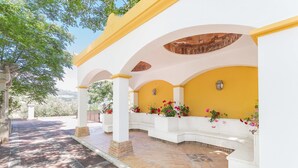 Un área de estar al aire libre cubierta con columnas blancas y paredes amarillas, con plantas en macetas y piso de baldosas.