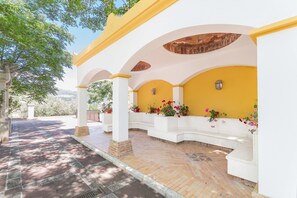 A covered outdoor area with white columns and yellow walls, featuring a brick floor and potted plants.