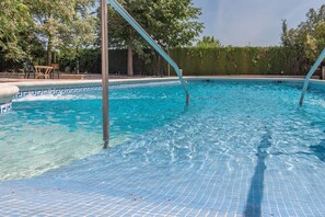 Una piscina cristallina con bordo piastrellato, circondata da alberi e un'area relax con sedie.