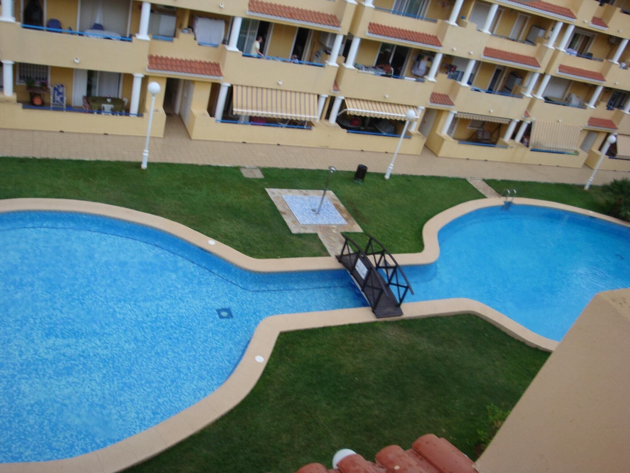 A swimming pool with a bridge, surrounded by a well-maintained lawn and a multi-story building with balconies.