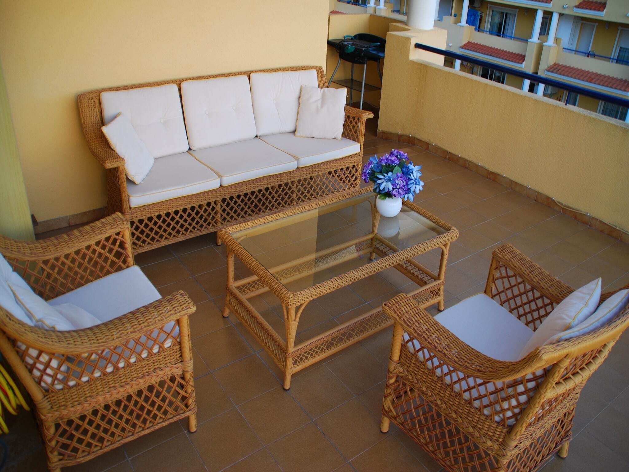 Un balcón con un sofá de mimbre, dos sillones de mimbre, una mesa de centro con tapa de vidrio y un jarrón con flores.