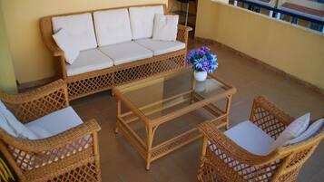 Un balcón con un sofá de mimbre, dos sillones de mimbre, una mesa de centro con tapa de vidrio y un jarrón con flores.