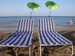 Twee gestreepte strandstoelen met groene parasols aan een zandstrand aan de zee.