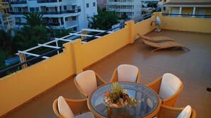 A round glass table with wicker chairs on a balcony, surrounded by yellow walls and a railing.