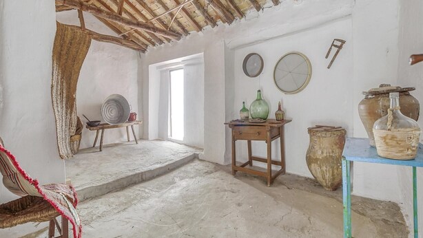 Una habitación rústica con techo de madera, una silla de fibras naturales, una mesita con cerámica y un jarrón grande en un estante.