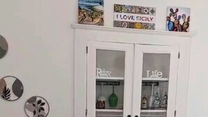 A white cabinet with glass doors, a small table with a vase of pink flowers, and wall decorations including a sign that says "I LOVE SICKLY". - Panoramic cottage 100 mt from the main square view Taormina and Calabria (Castelmola)