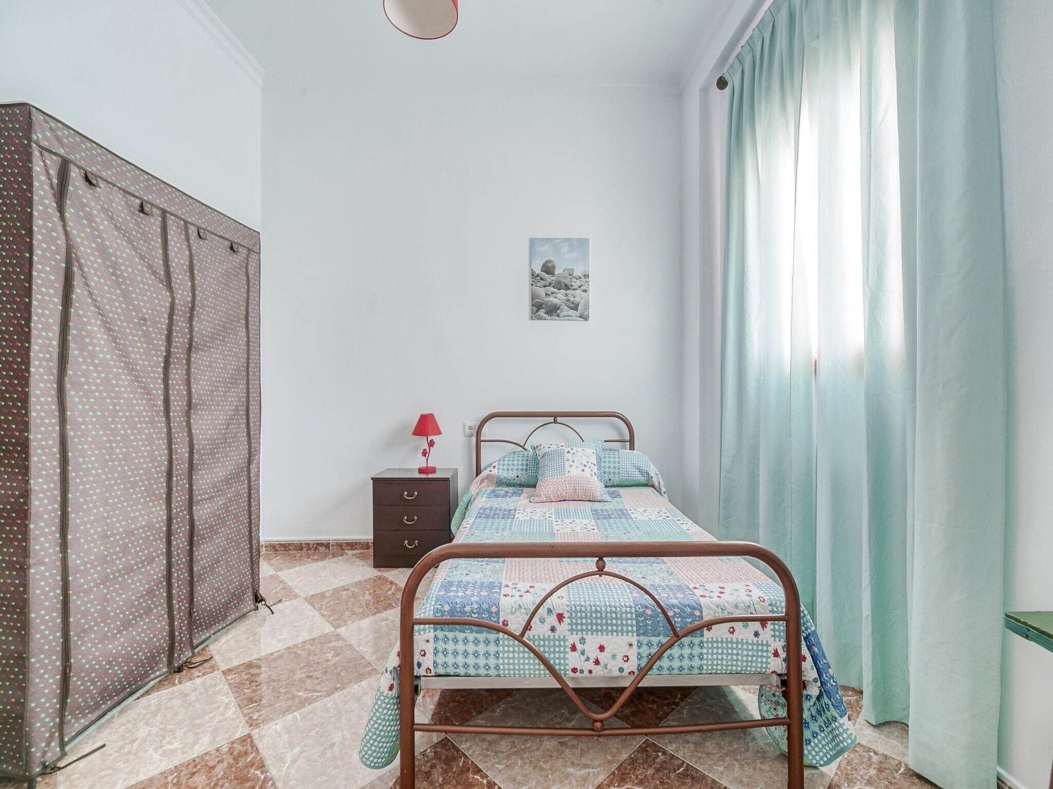 Une chambre à coucher avec un lit en métal, une petite table de chevet avec une lampe, un tableau accroché au mur et une fenêtre avec des rideaux.