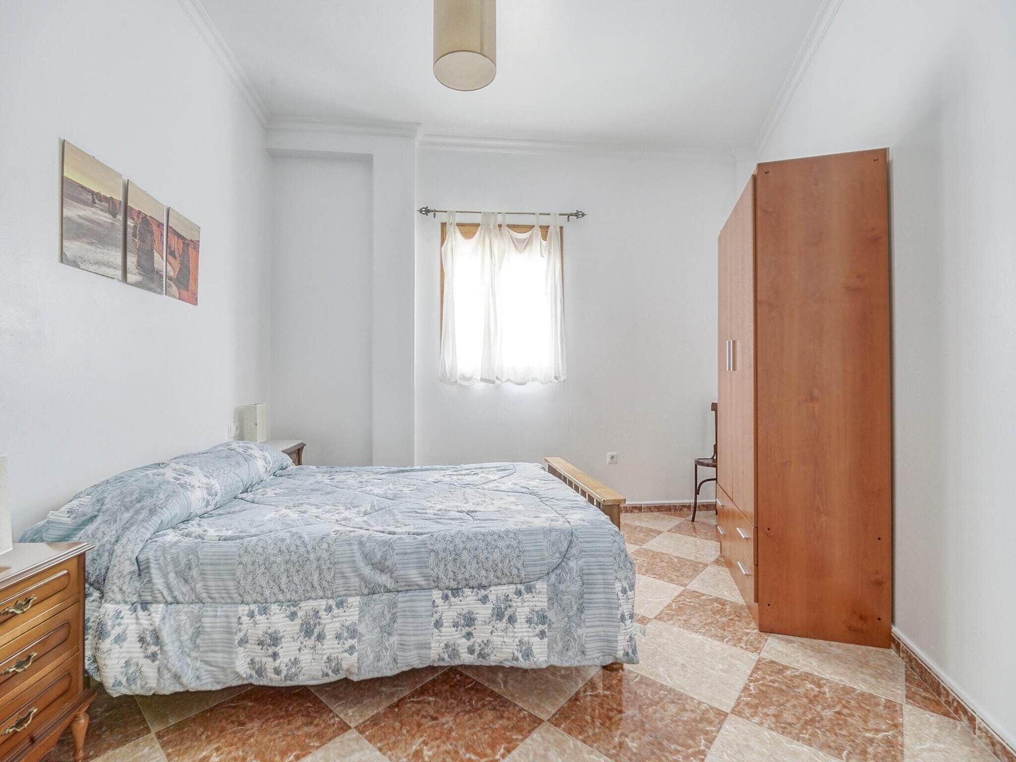 Una camera da letto con un letto, un armadio in legno, una panca, una finestra con tende e un quadro appeso al muro.