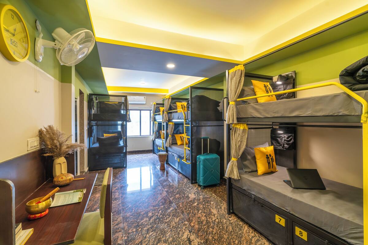 Une chambre d’hôtel compacte avec des lits superposés, un bureau avec un ordinateur portable, un ventilateur et une horloge murale.