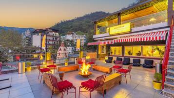 A rooftop bar with a fire pit, red and white striped awnings, and a view of the surrounding buildings and hills.
