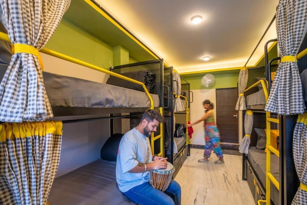 A dormitory room with bunk beds, a person playing a drum, and another person walking.