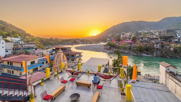 A terrace with outdoor seating, a view of a river, and buildings in the background. - The Hosteller Rishikesh, Ganges (RISHIKESH)
