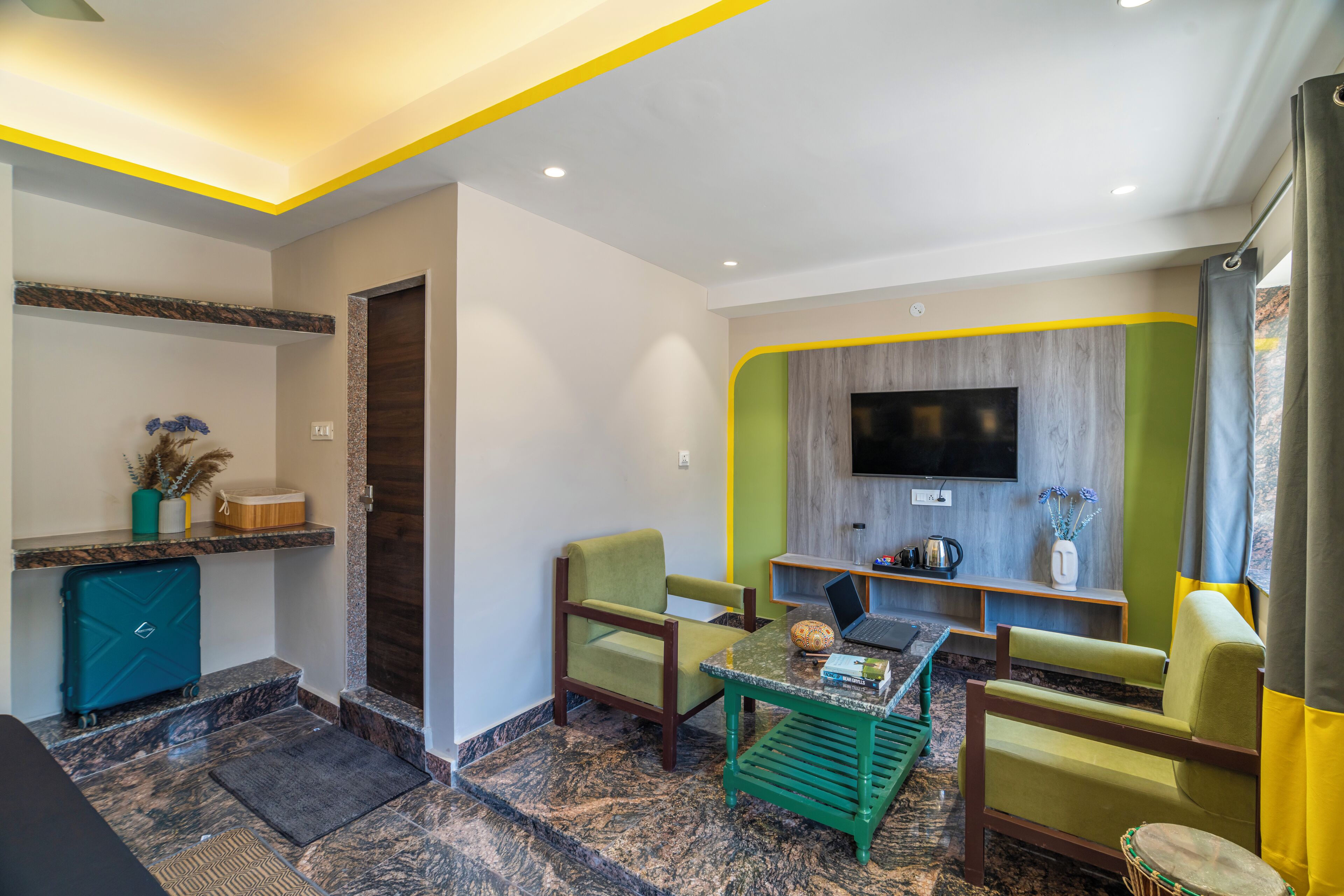 Un salón moderno con un televisor de pantalla plana montado en la pared, dos sillones verdes, una mesa de centro y un ordenador portátil sobre una mesita de noche de madera.