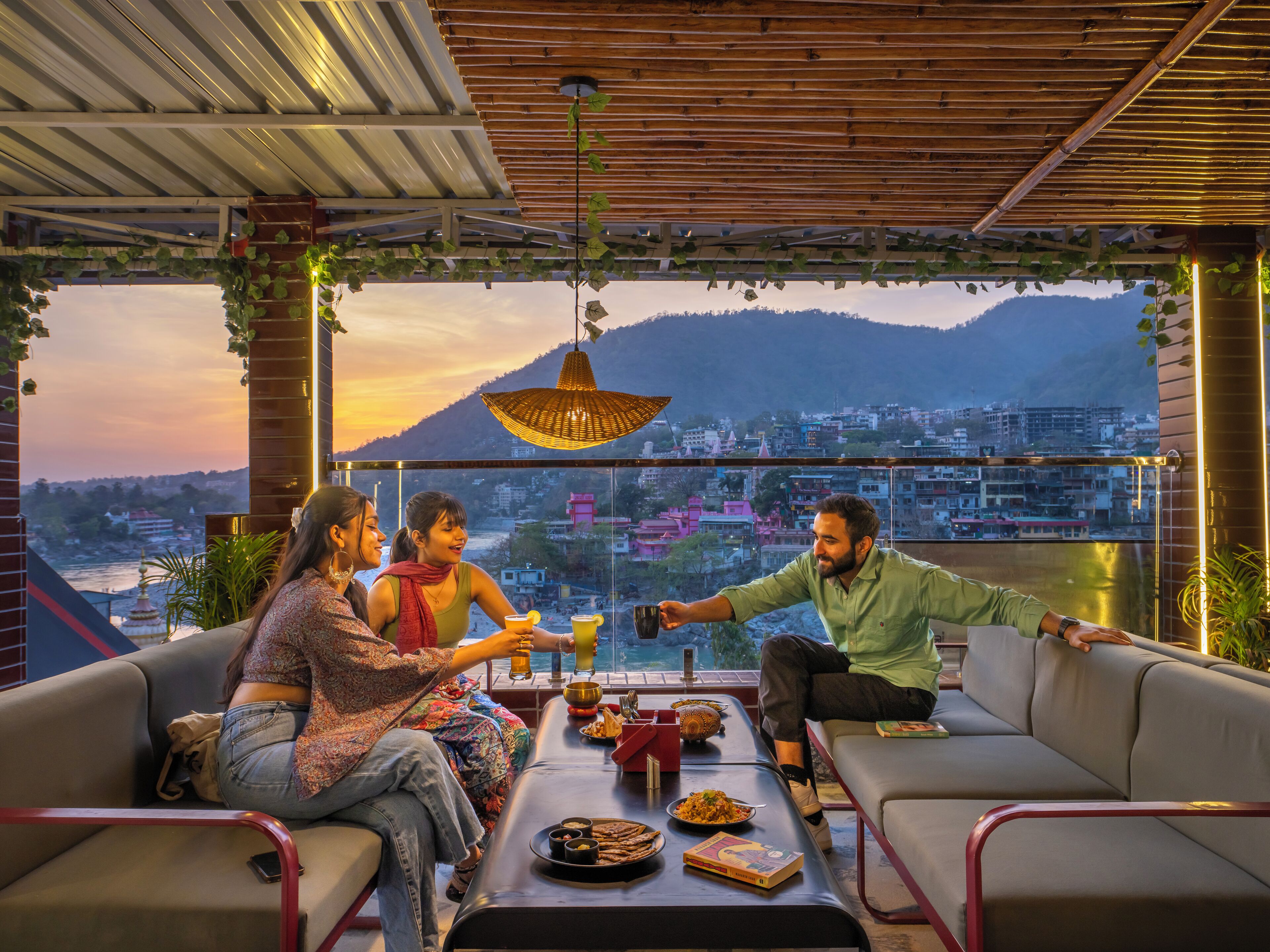 Una terraza con vistas a un paisaje urbano y montañas, con una mesa llena de comida y bebidas, y disposición de asientos.