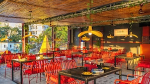 An outdoor dining area with red chairs and tables, bamboo ceiling, and hanging yellow lamps. - The Hosteller Rishikesh, Ganges (RISHIKESH)