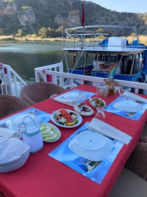 A dining table set with plates, glasses, and condiments on a boat with a view of a rocky landscape and a building in the background.