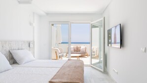 A modern hotel room with two beds, a large sliding glass door leading to a balcony with a view of the sea, and a flat-screen TV mounted on the wall.