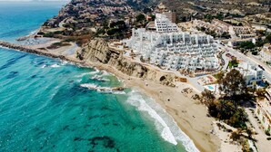 An aerial view of a coastal town with white buildings, a beach, and mountains in the background.
