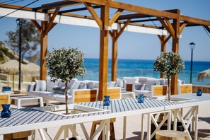 A sunlit outdoor seating area with white lounge chairs and tables, a wooden pergola, and a view of the sea.