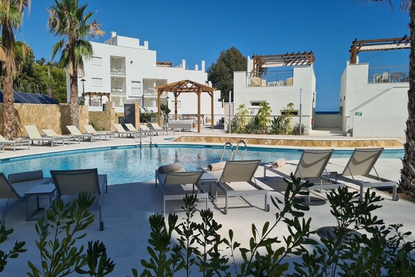 A pool area with lounge chairs, a wooden pergola, and a view of the sea.