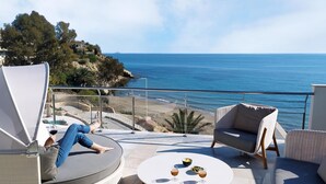 A balcony with a view of the sea, a round table with drinks, and a reclining chair.