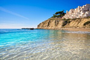 Un paysage côtier avec une eau turquoise cristalline, une plage de sable et une falaise sur laquelle se trouvent des bâtiments.