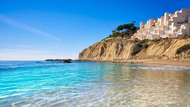 A coastal scene with clear turquoise waters, a sandy beach, and a cliff with buildings.