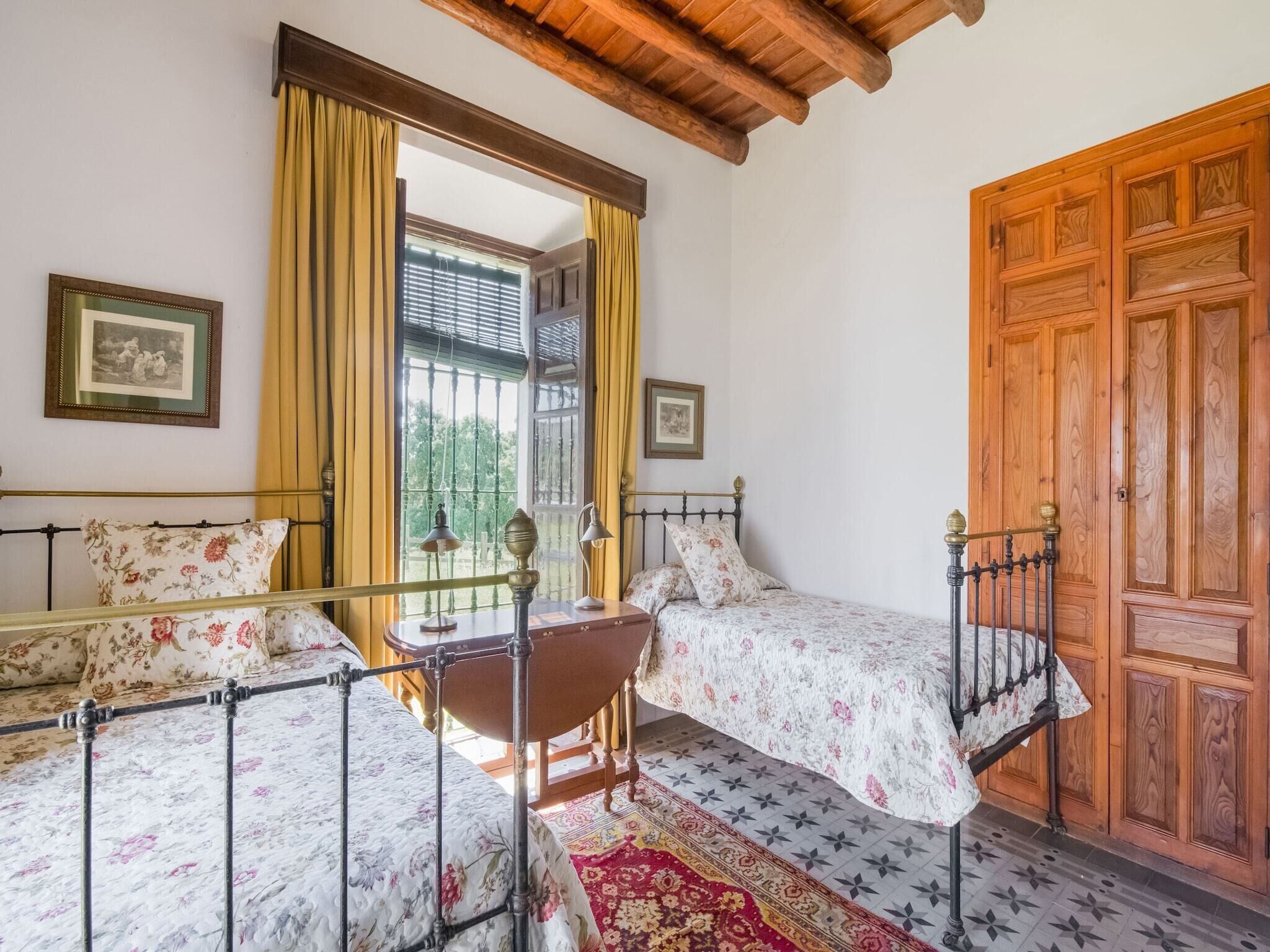 A bedroom with two single beds, a wooden wardrobe, a window with curtains, and a small table with a lamp.